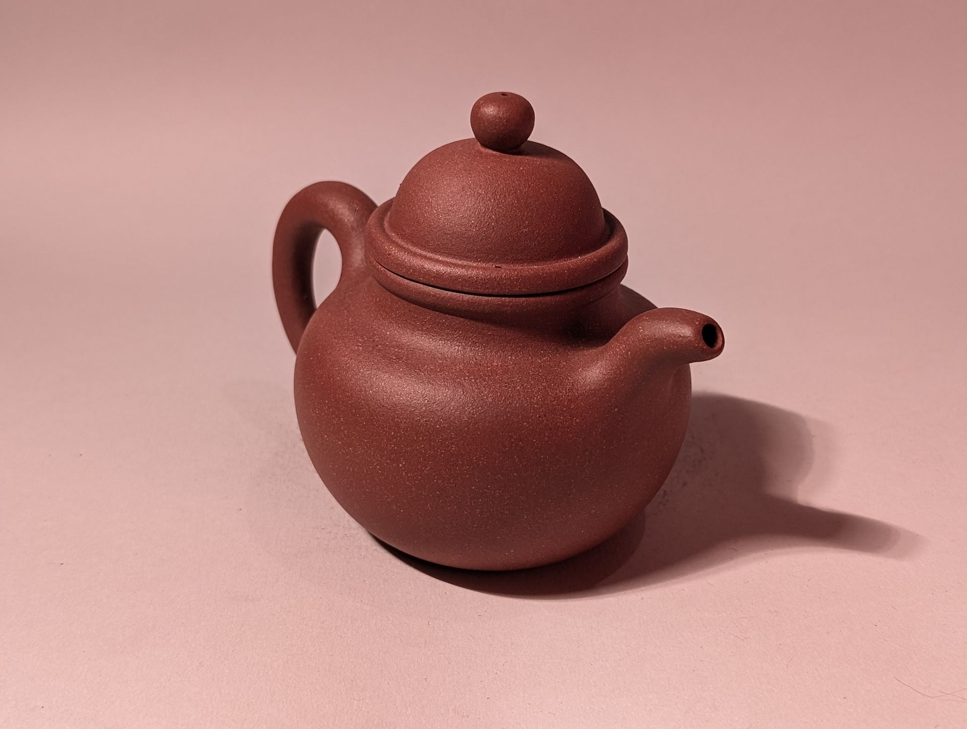 Small brown teapot with a lid and handle on a pink background