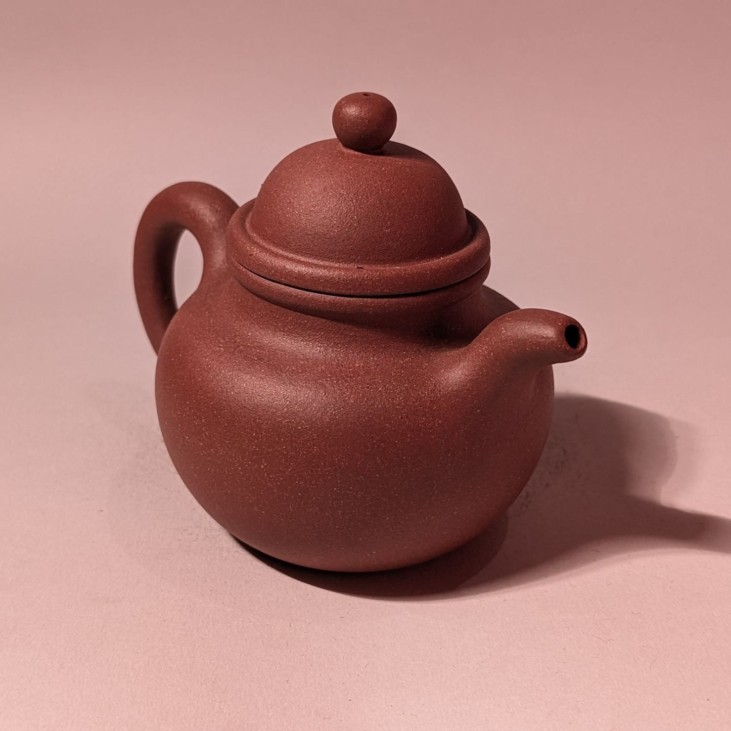 Small brown ceramic teapot with a round lid and curved handle on a pink surface.