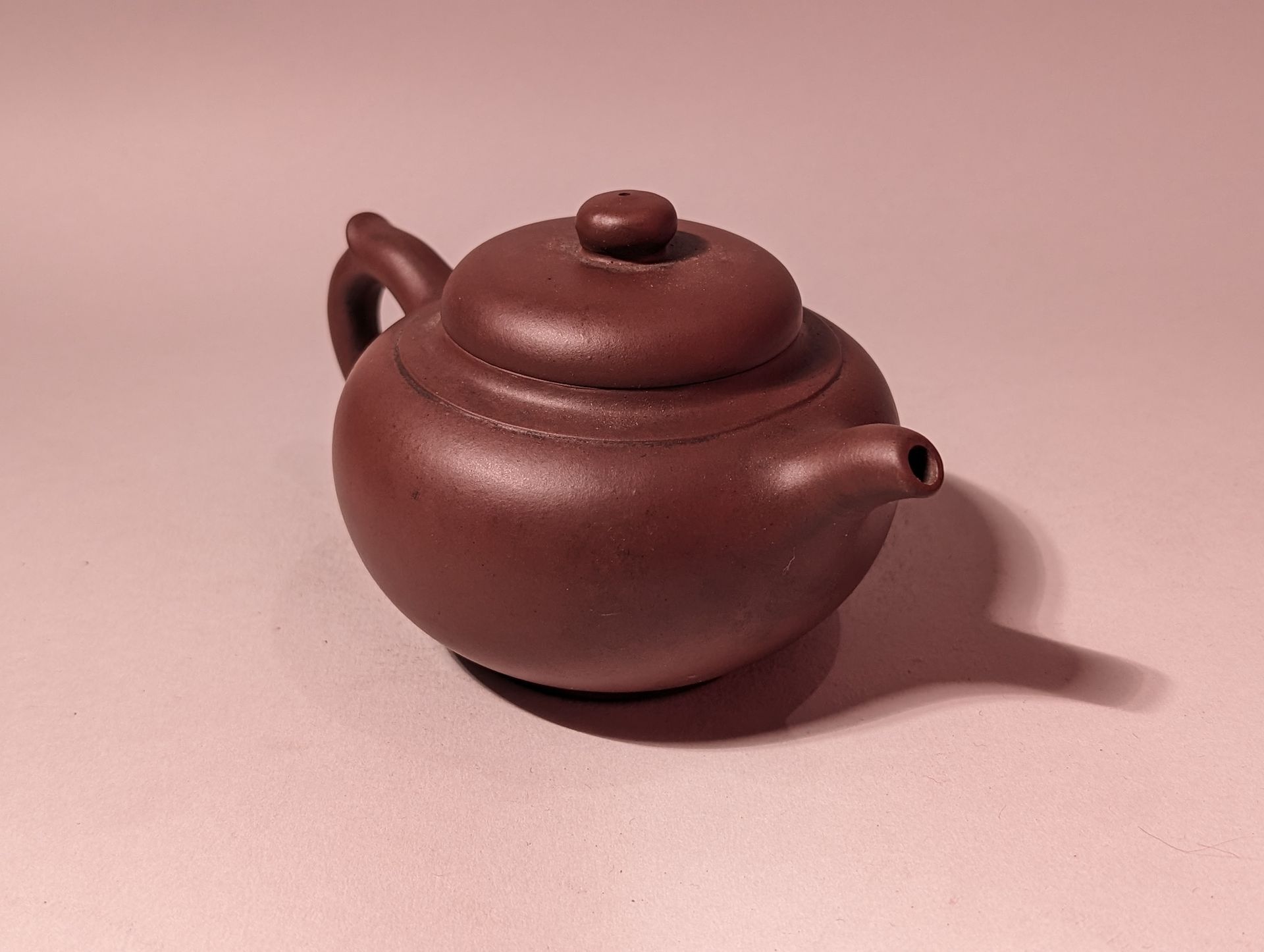 Small brown clay teapot with a lid and curved handle on a pink background