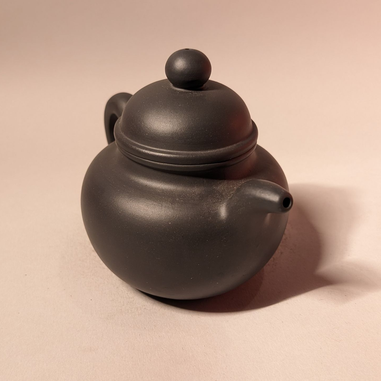 Small dark ceramic teapot with round lid and short spout on a plain beige background