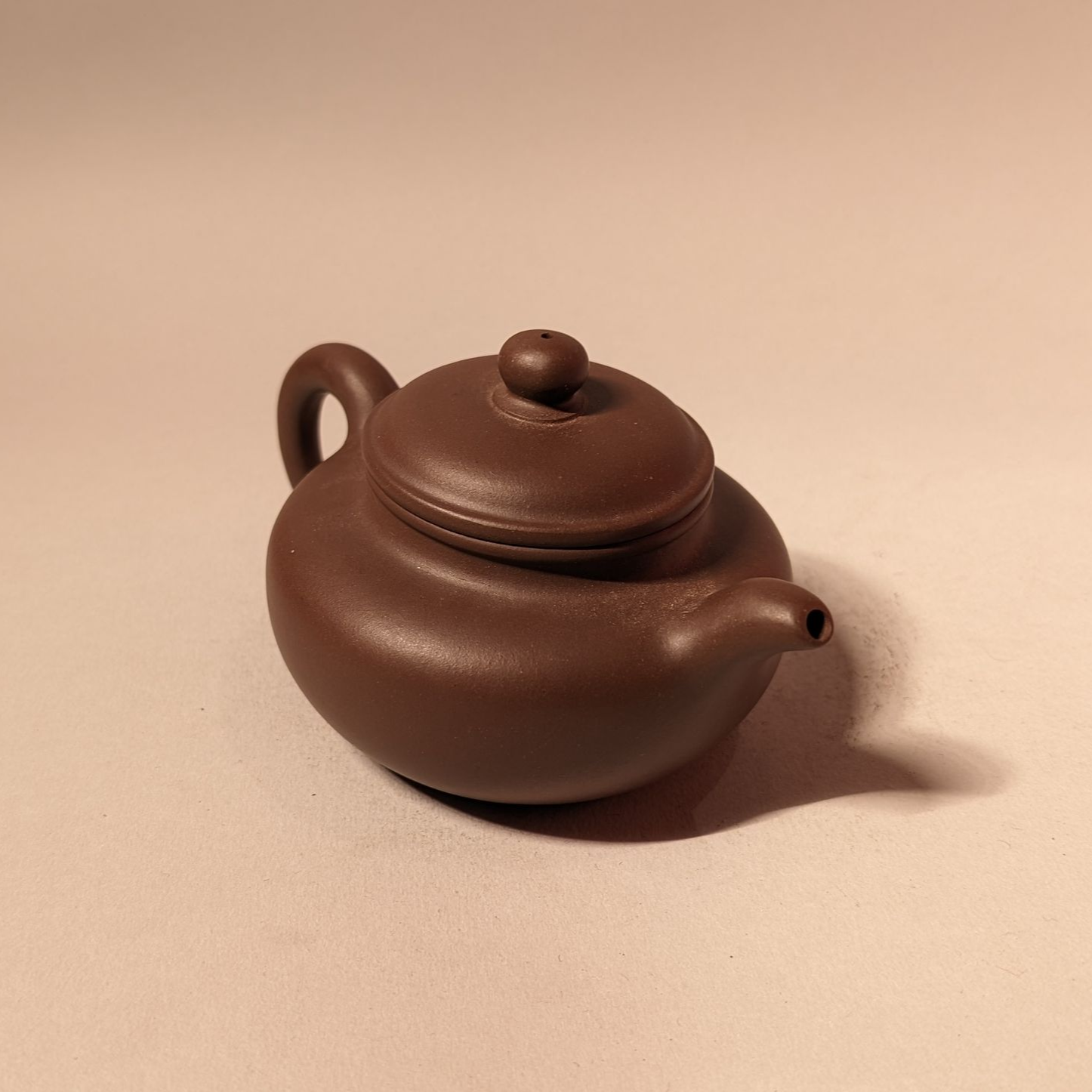 Small brown ceramic teapot with lid and spout on a beige surface