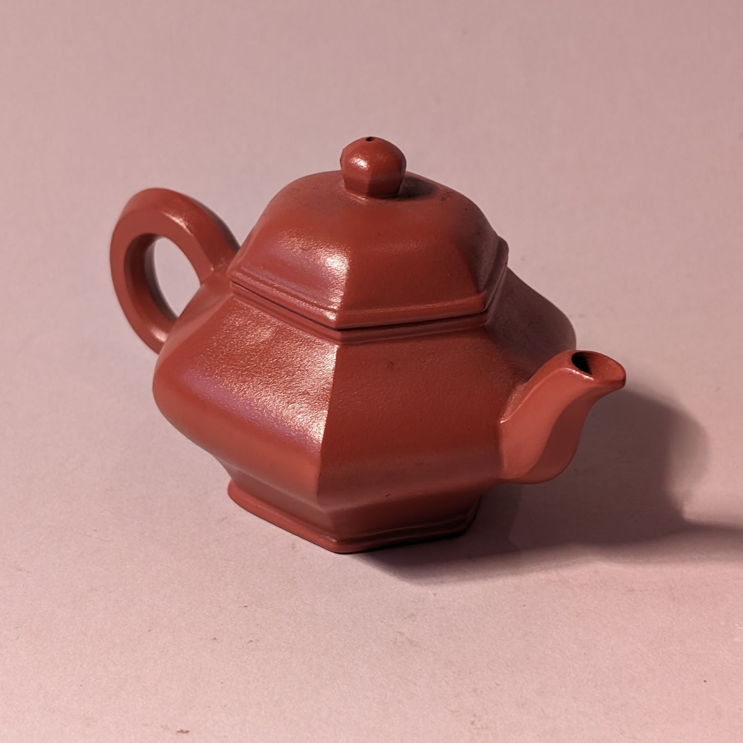 Small reddish-brown ceramic teapot with a lid on a pale pink background