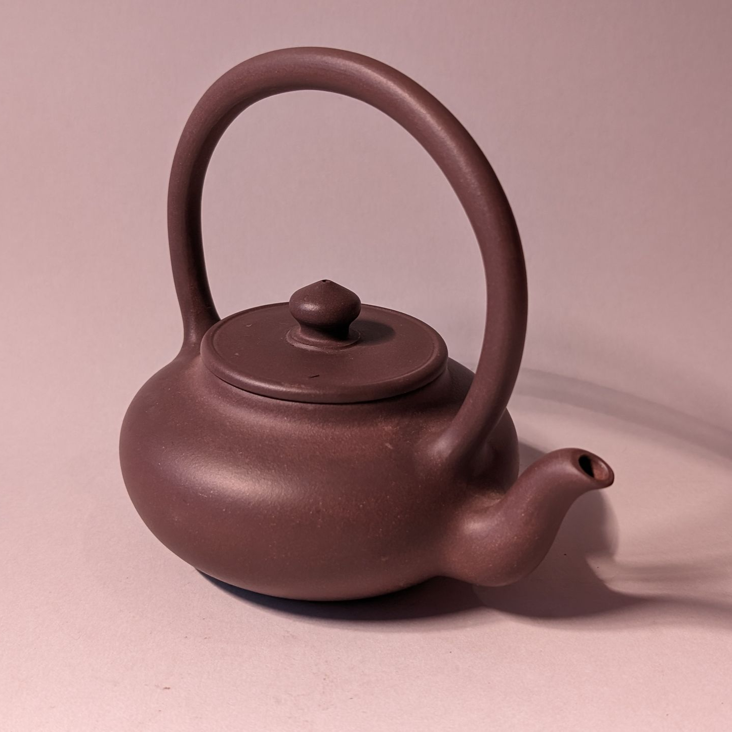 Small brown ceramic teapot with a tall handle and round lid on a pink background