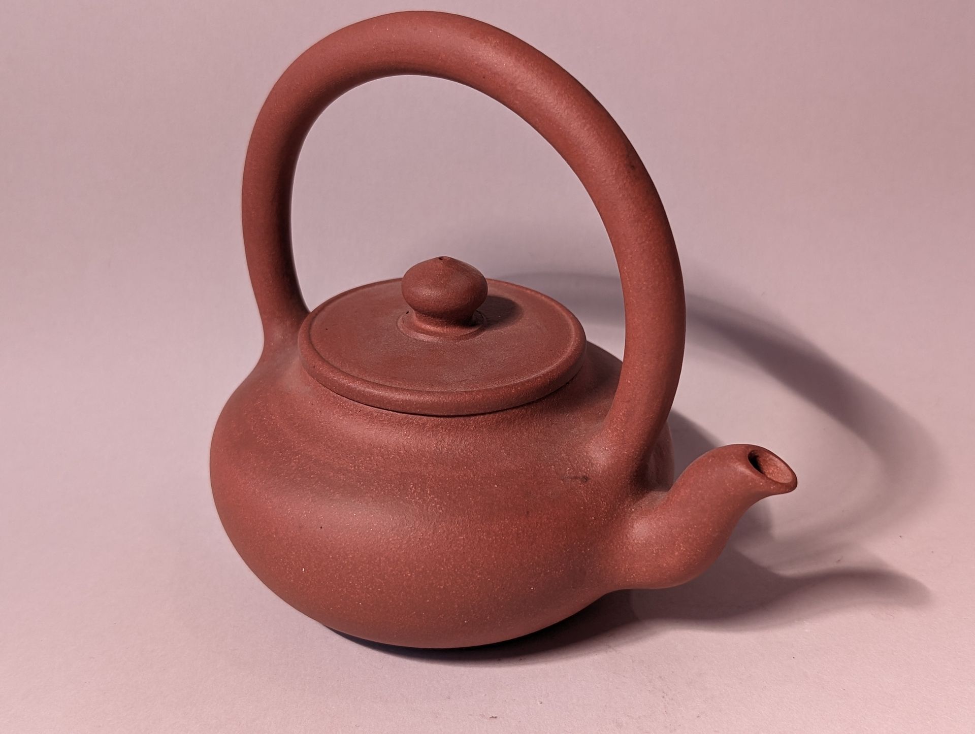 Brown clay teapot with a tall arched handle and short spout on a pink background
