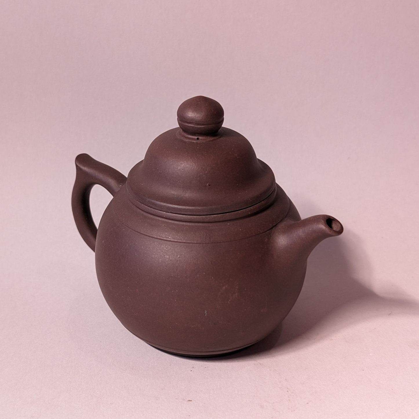 Brown clay teapot with lid and curved handle on a pale pink background