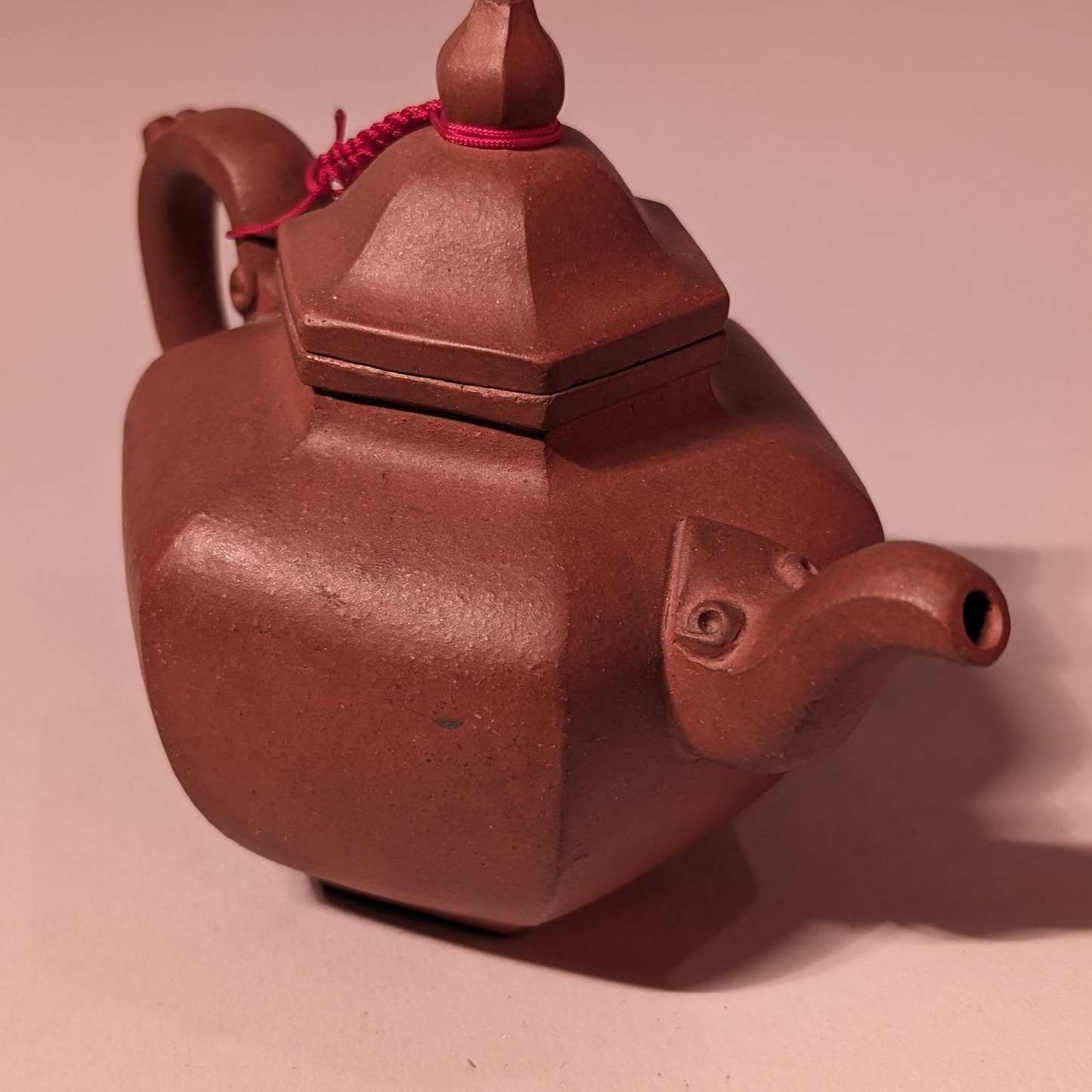 Small brown ceramic teapot with a square body and red cord, on a pink surface.