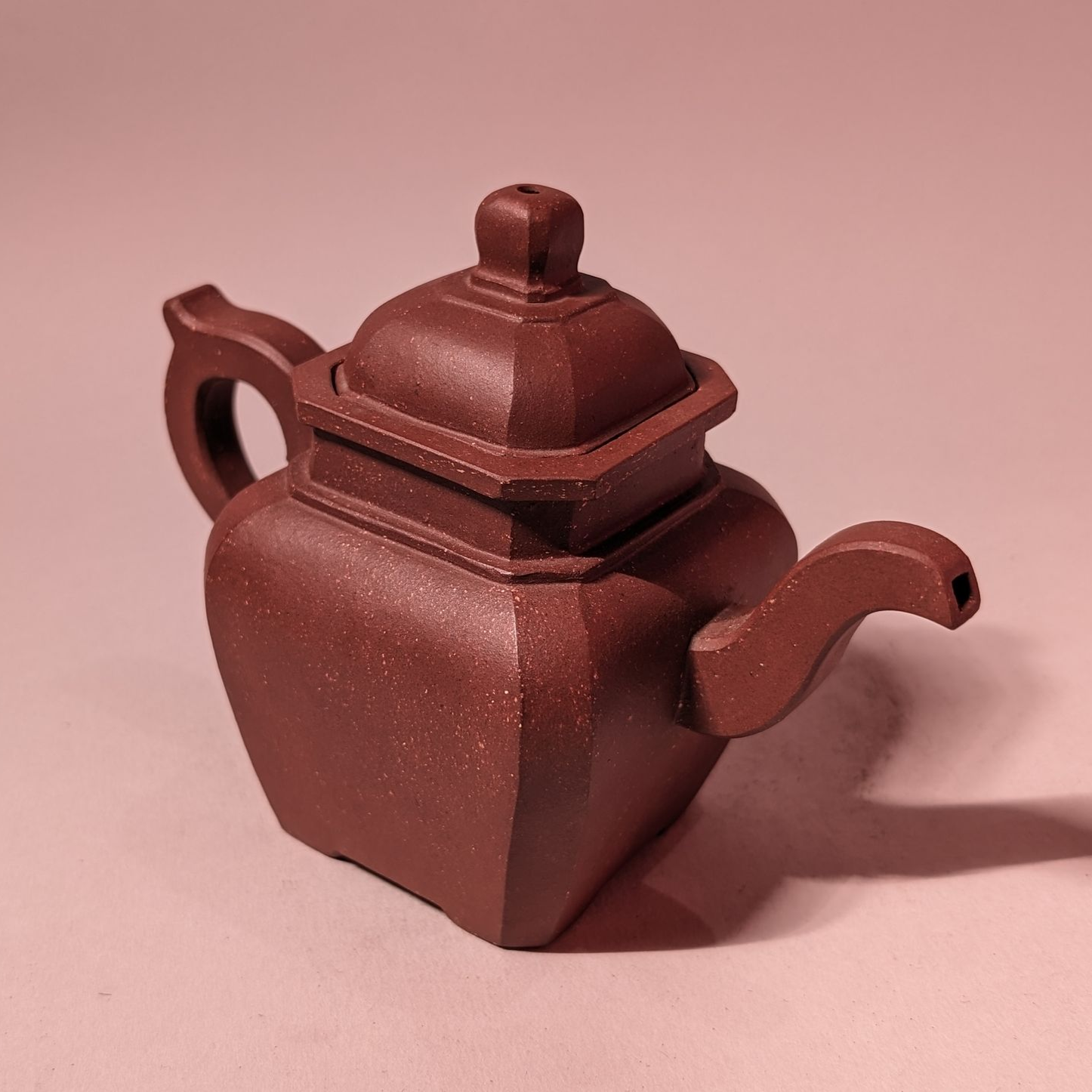 Brown ceramic teapot with a square body and curved handle on a pink background