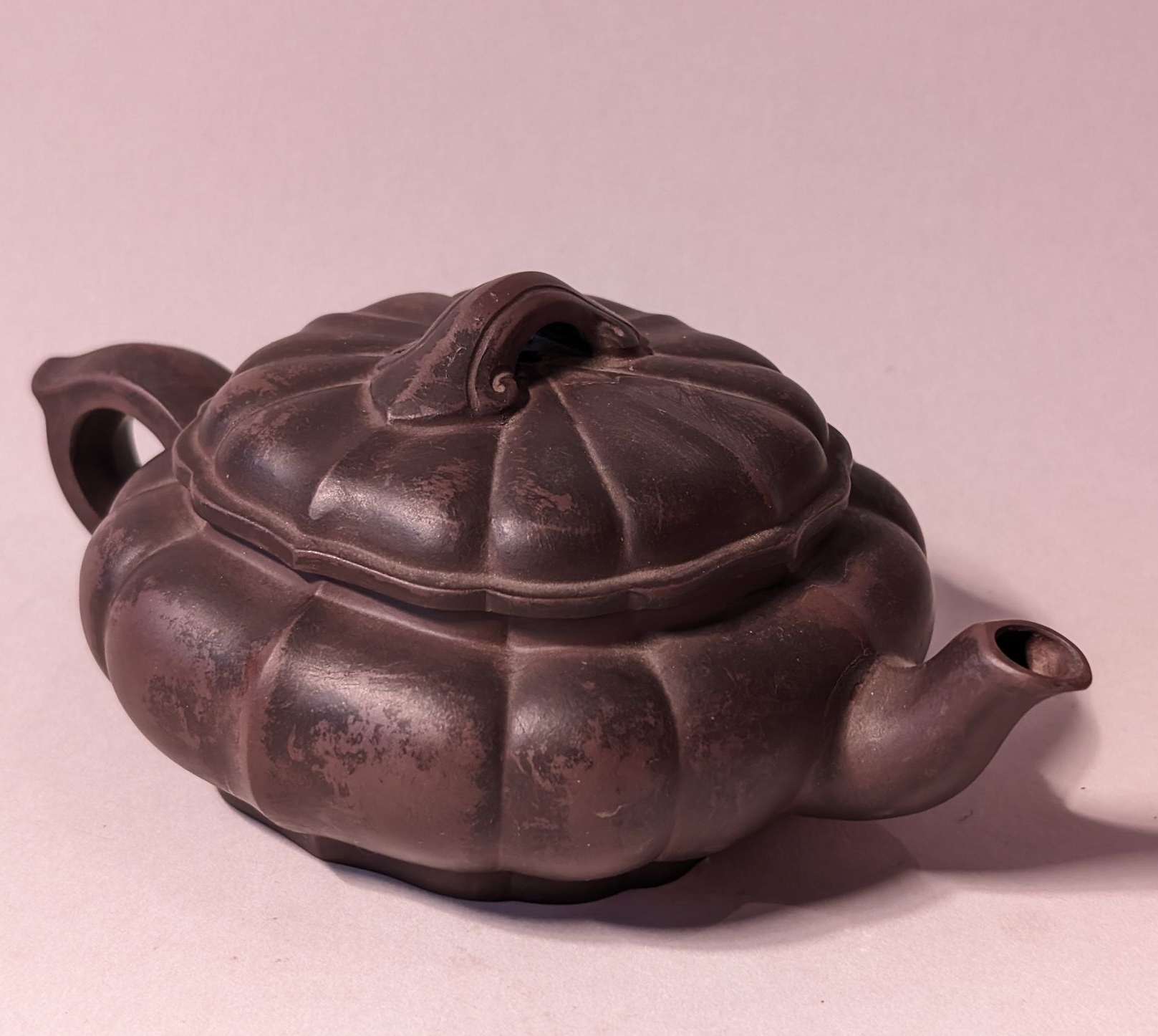 Dark brown ceramic teapot with a rounded lid and curved spout on a light pink background