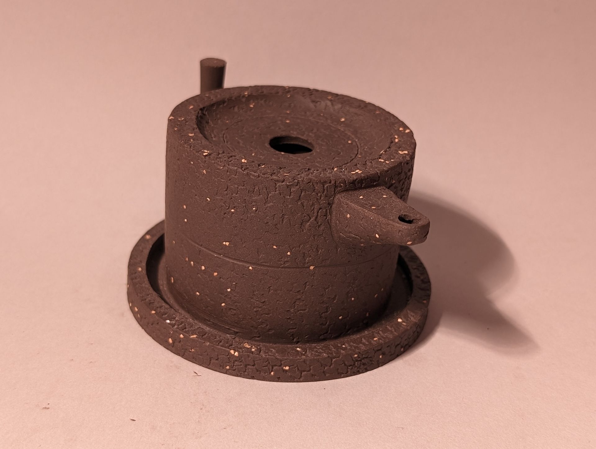 Brown metal cylindrical device with a round base, central hole, and small side nozzle on a pink background