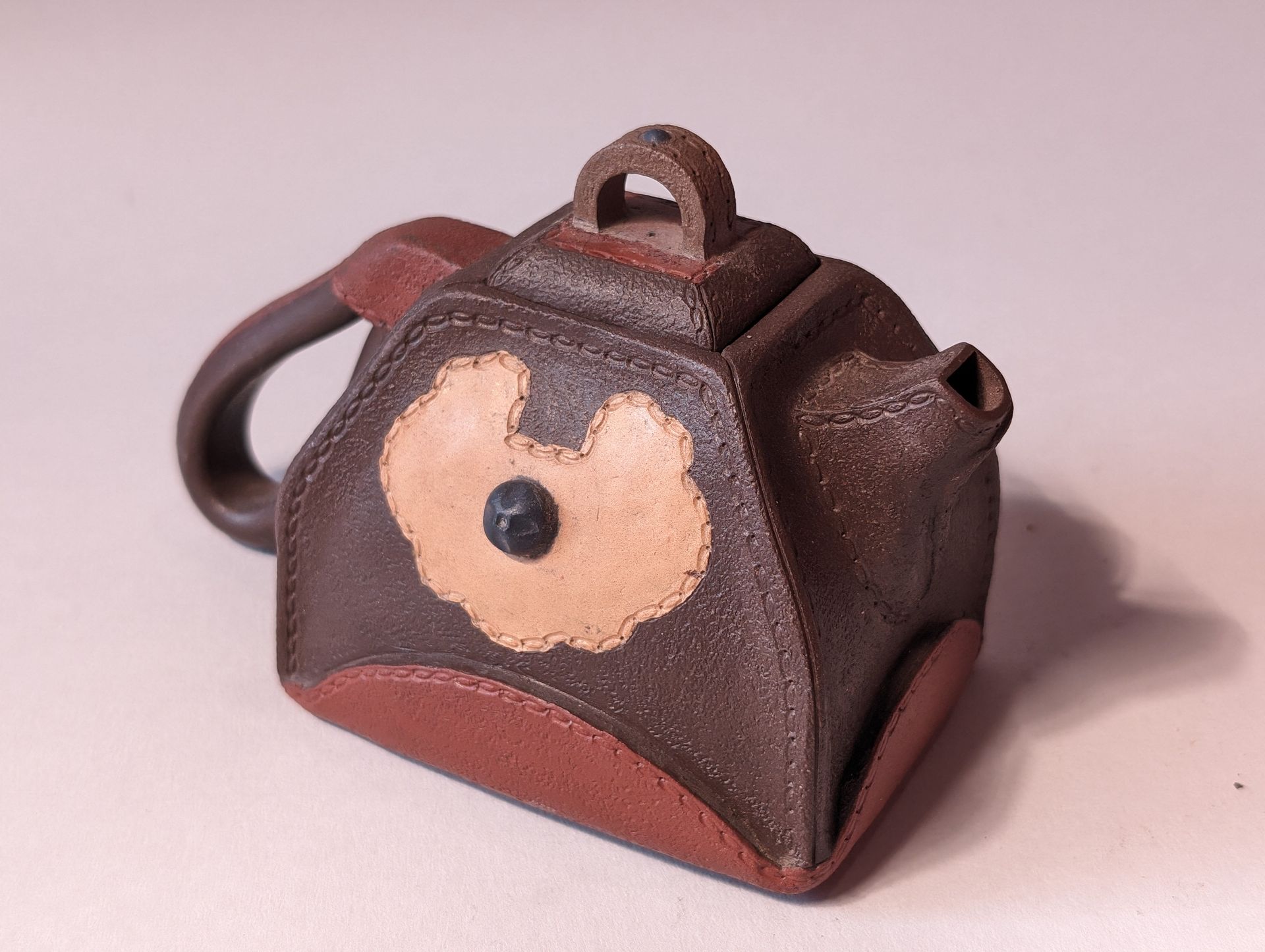 Small brown decorative teapot-shaped ceramic container with a handle and floral face design