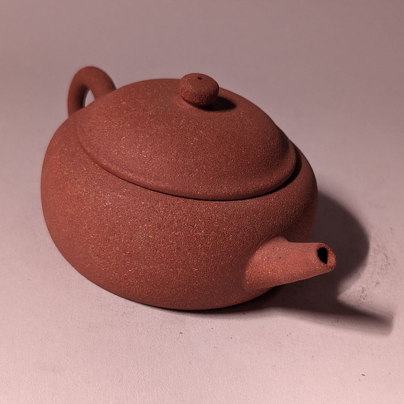 Small reddish-brown ceramic teapot with lid and handle on a pink background