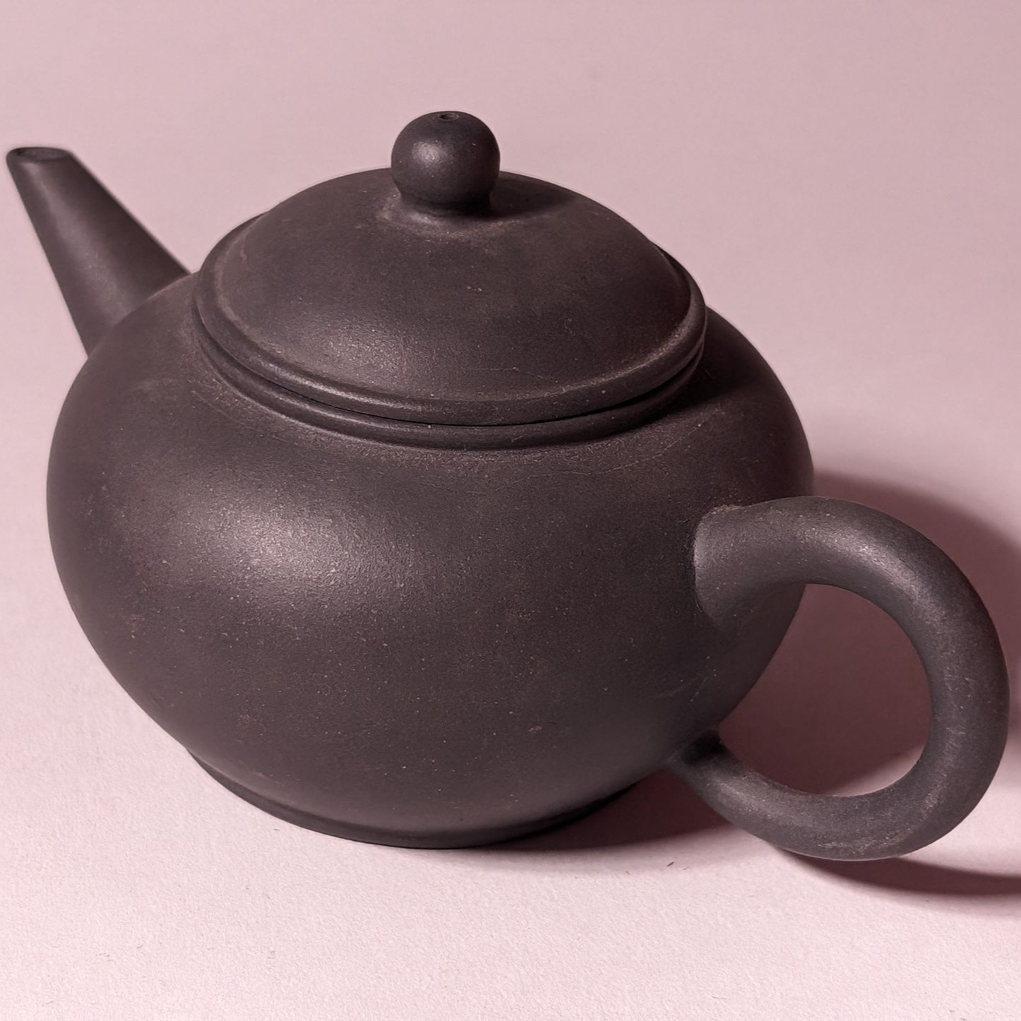 Dark gray ceramic teapot with lid and round handle on a light pink background