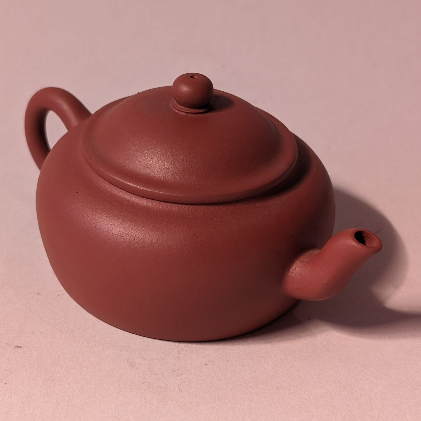 Small reddish-brown teapot with lid and curved handle on a pink background