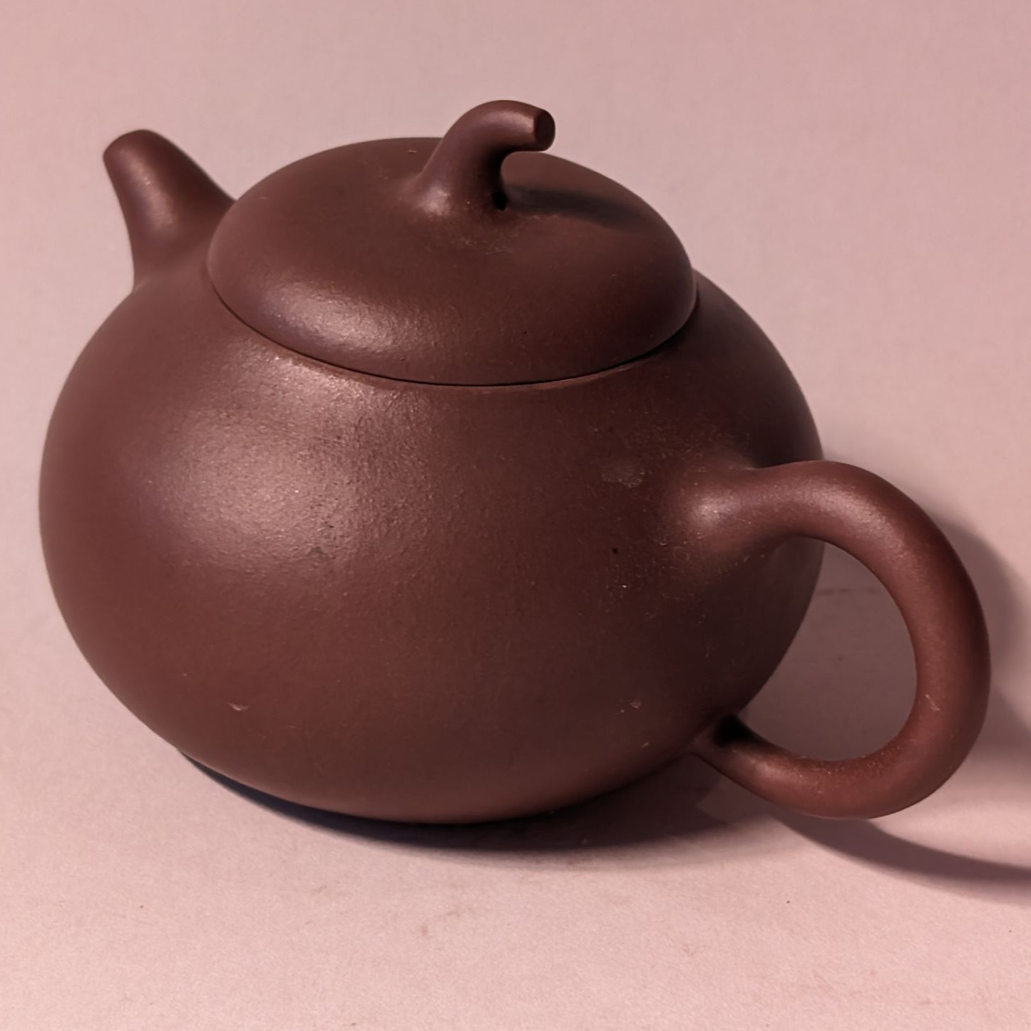 Brown ceramic teapot with round body and curved handle on a pink surface