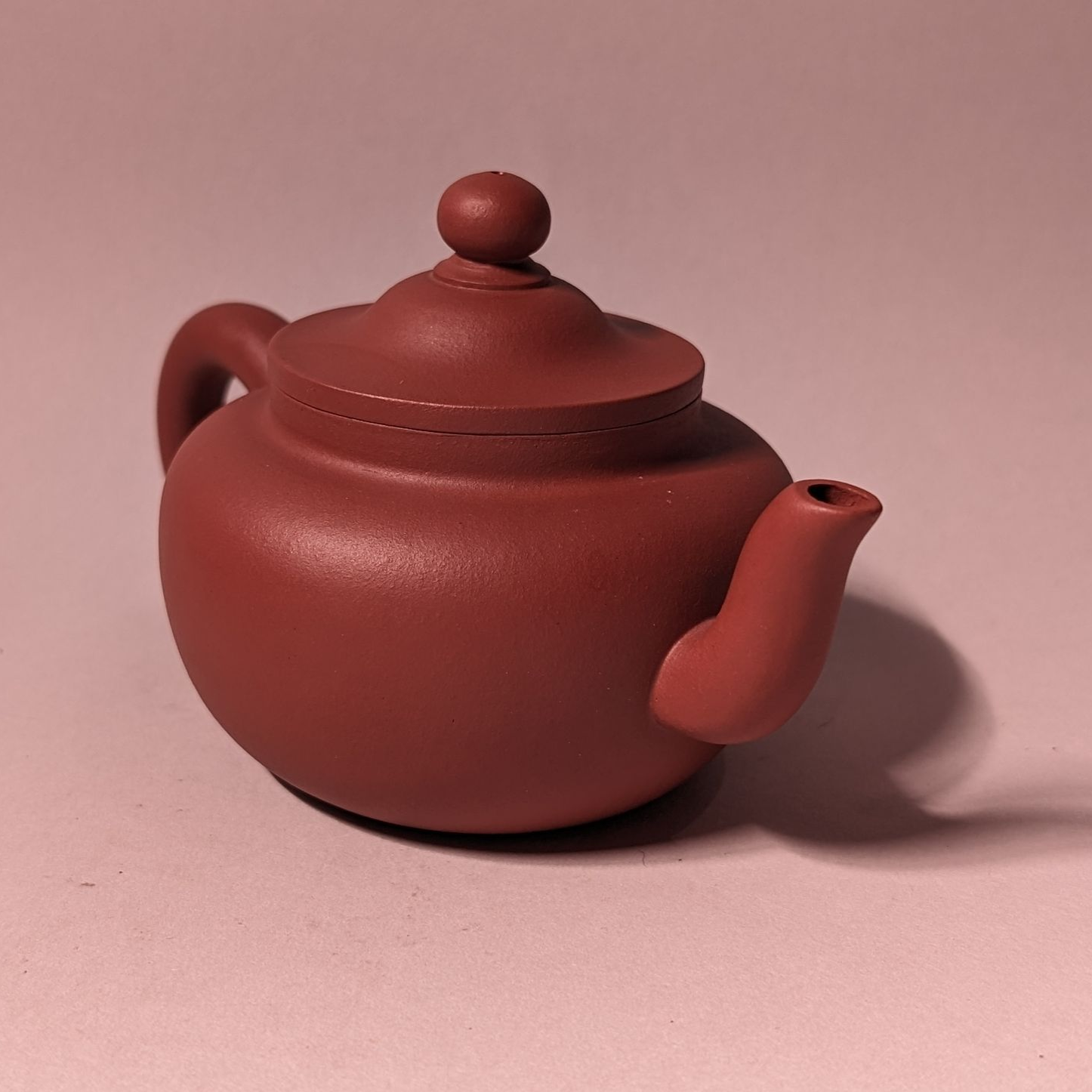 Small reddish-brown ceramic teapot with lid and curved spout on a pink surface