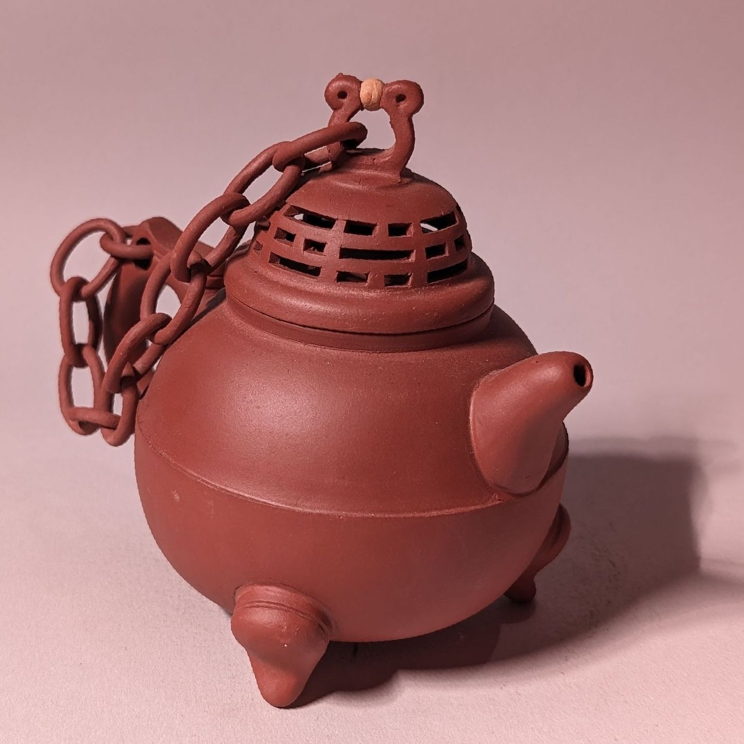 Small reddish-brown ceramic teapot with a chain, rounded body, and lid, shown on a pink background