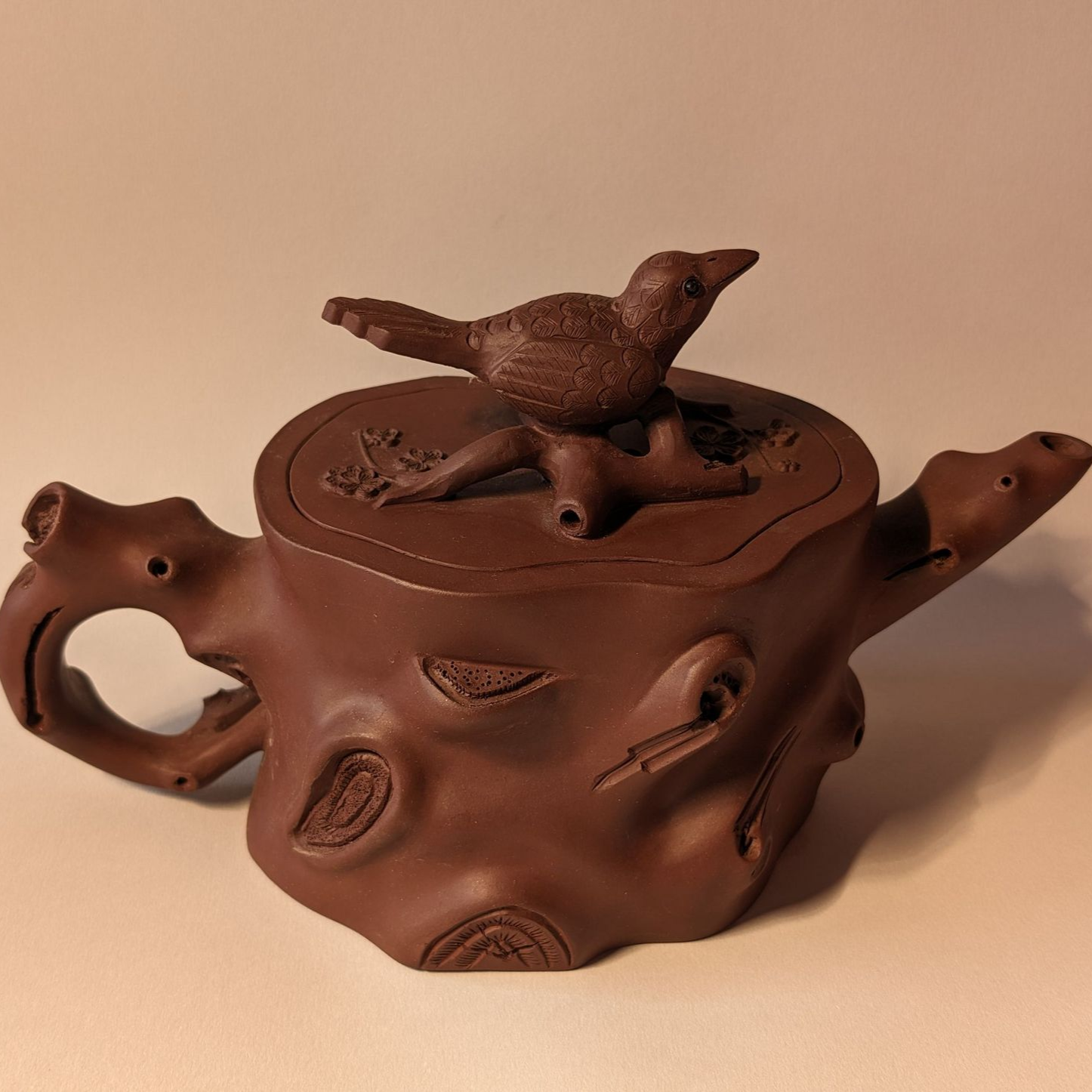 Brown ceramic teapot with a bird-shaped lid and twig-like handle on a plain beige background