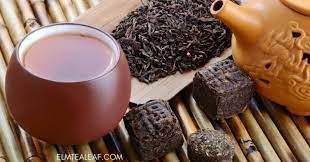 Glass of tea with loose tea leaves, tea bricks, and a small clay teapot on bamboo mat