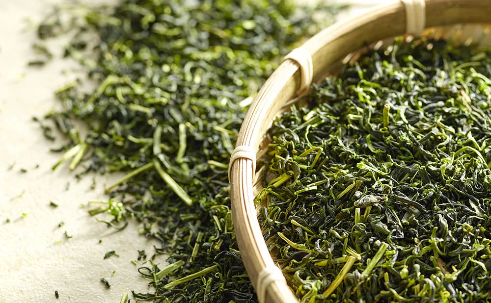 Bamboo tray with loose green tea leaves scattered on a light surface