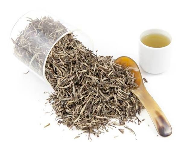 Loose dried tea leaves spilled from a tipped glass jar beside a spoon and a cup of tea.