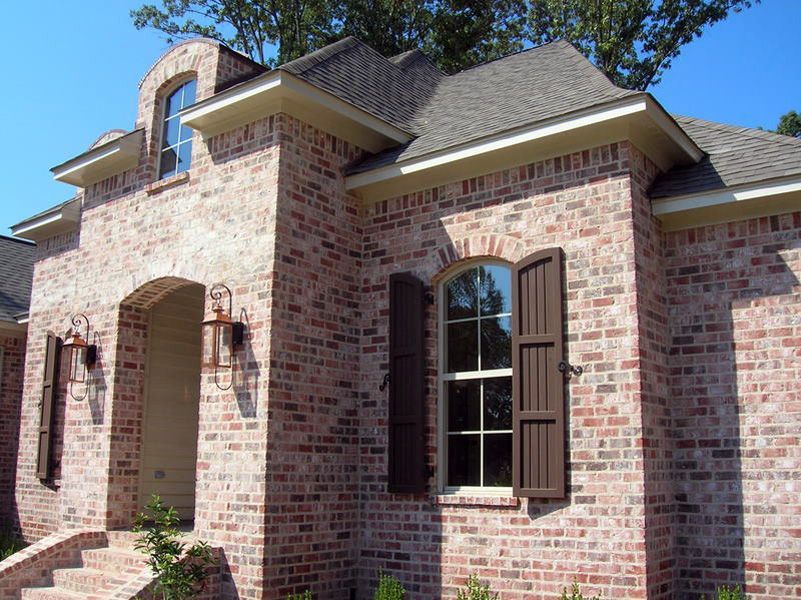 Brick house with arched doorway, dark brown shutters, and beige trim against a blue sky.