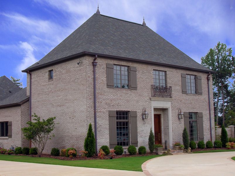 Two-story brick house with gray roof, brown shutters, and manicured landscaping, on a sunny day.