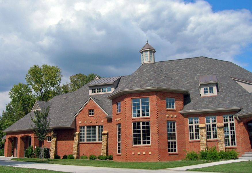 Red brick building with gray shingle roof, arched entryway, and multiple windows against a cloudy sky.