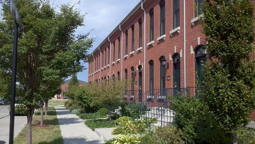 Row of red brick townhouses with green front doors, sidewalk, and trees.