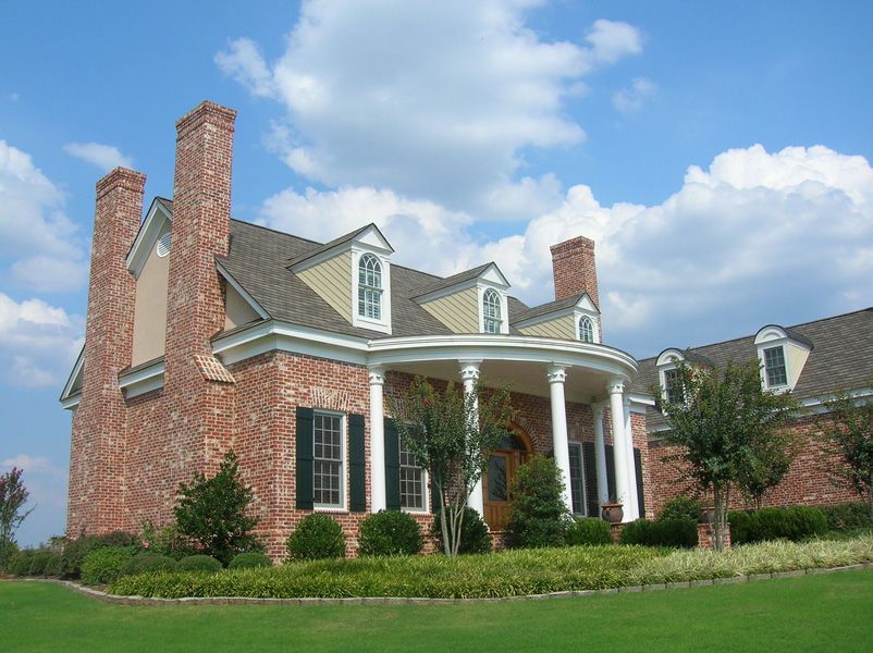 Red brick house with white columns, green lawn, and blue sky with clouds.
