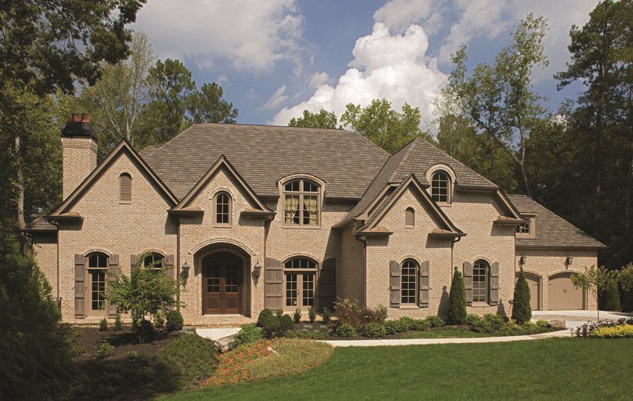 Brick house with brown roof, shutters, and arched windows, set in a yard with trees and blue sky.