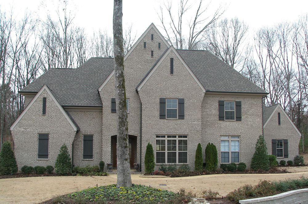 Beige brick house with multiple gables and gray roof, in a wooded setting.