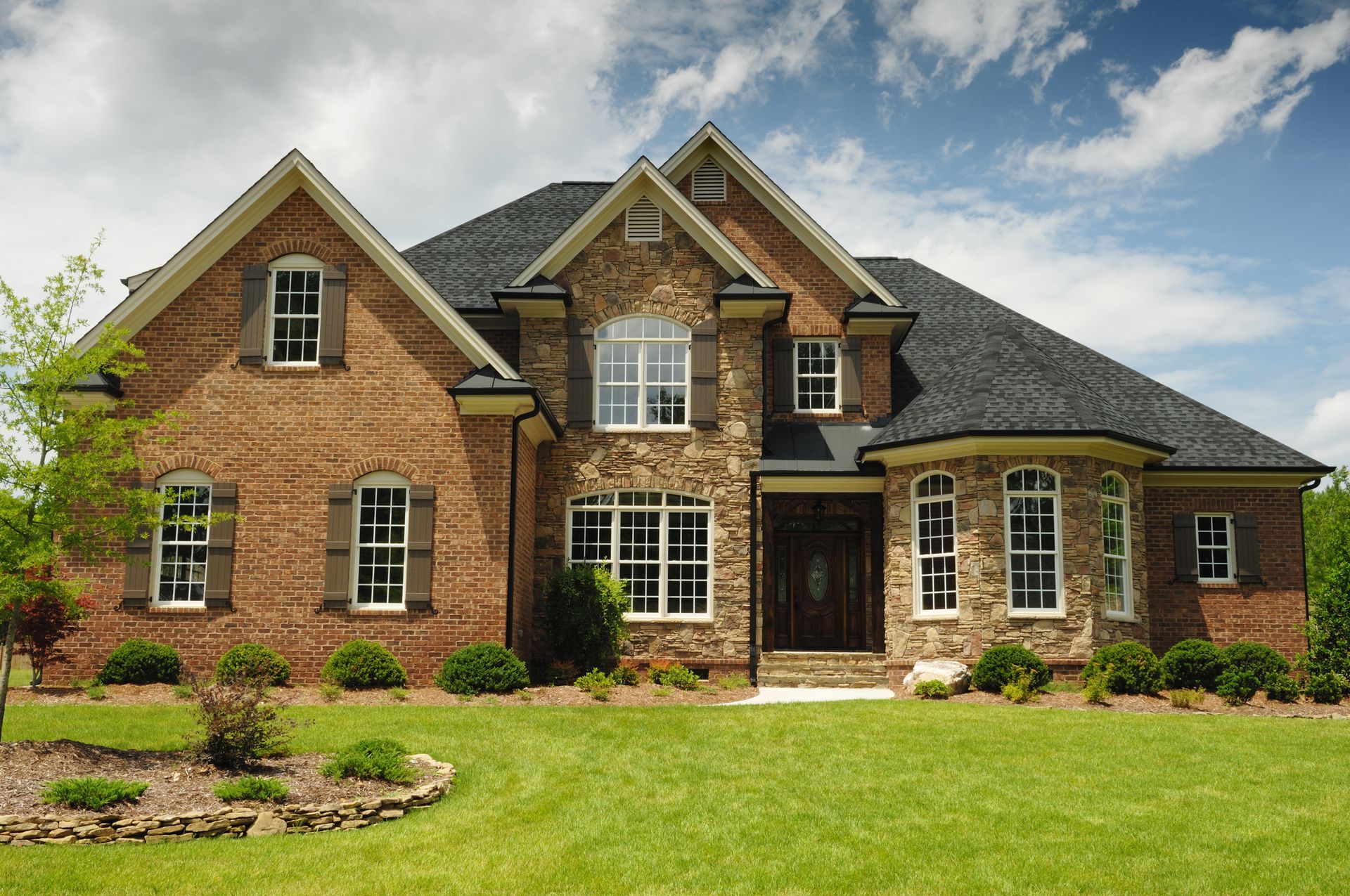 Brick and stone two-story house with green lawn and blue sky.