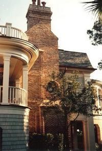 Brick chimney on a house with a shingled roof, next to a curved porch with a white railing.