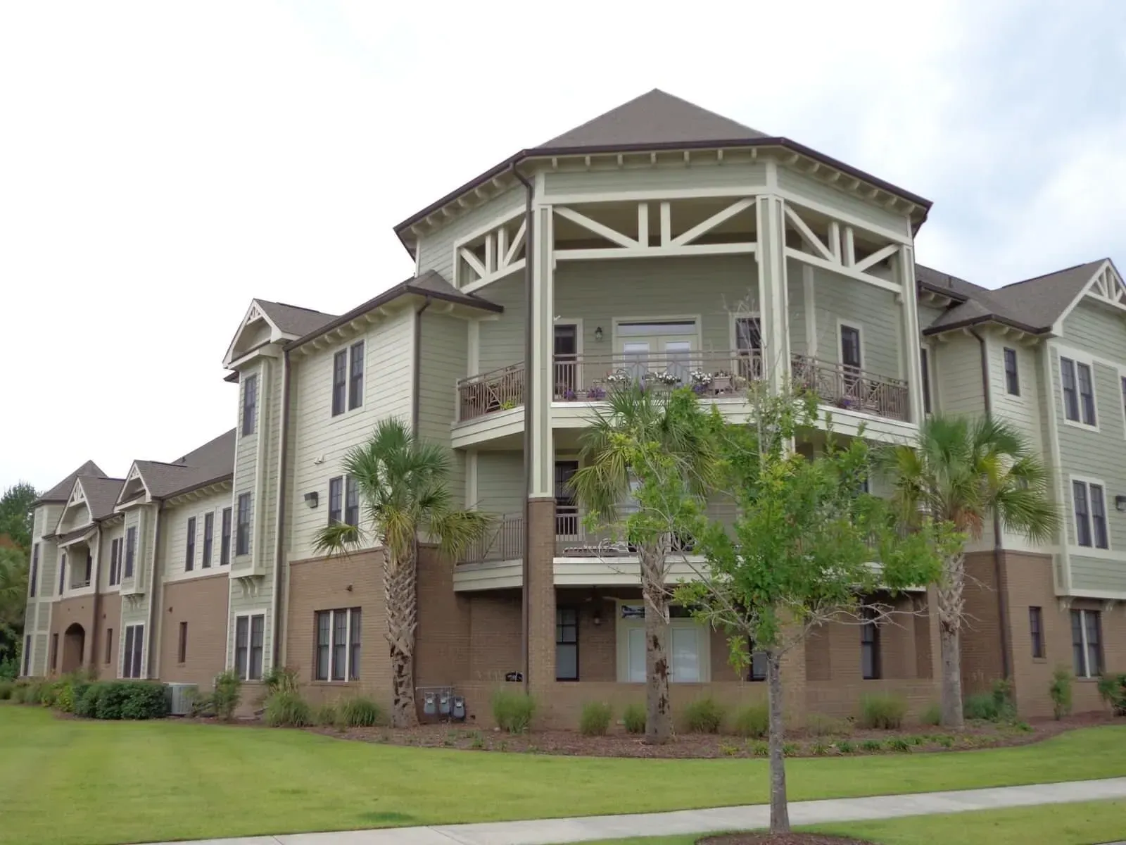 Multi-story apartment building with light green siding and brown brick base, with balconies and palm trees.