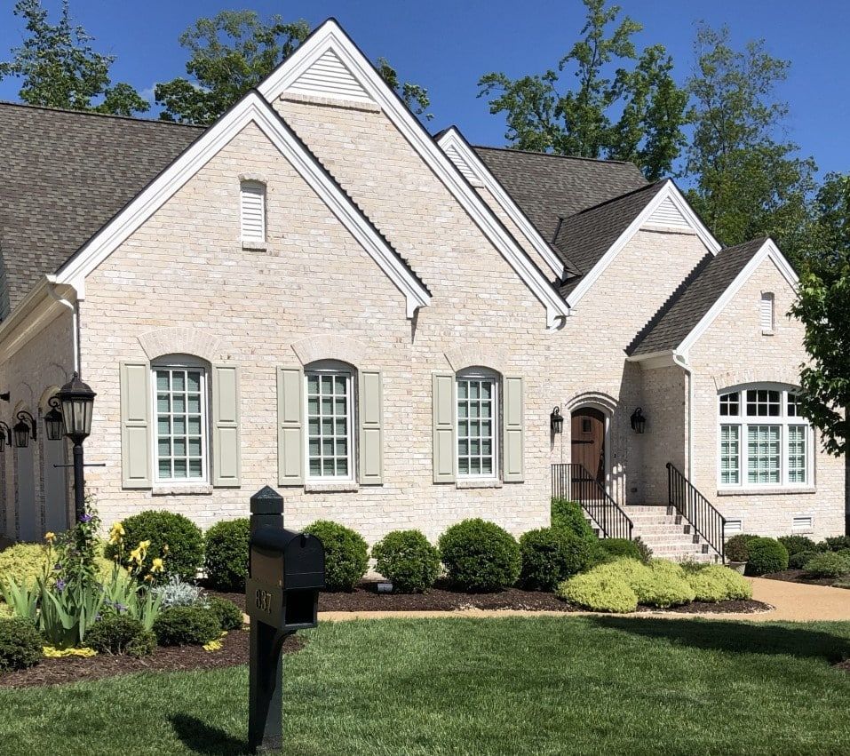 Cream-colored brick house with light green shutters and a dark roof. Lush green lawn and bushes in front.