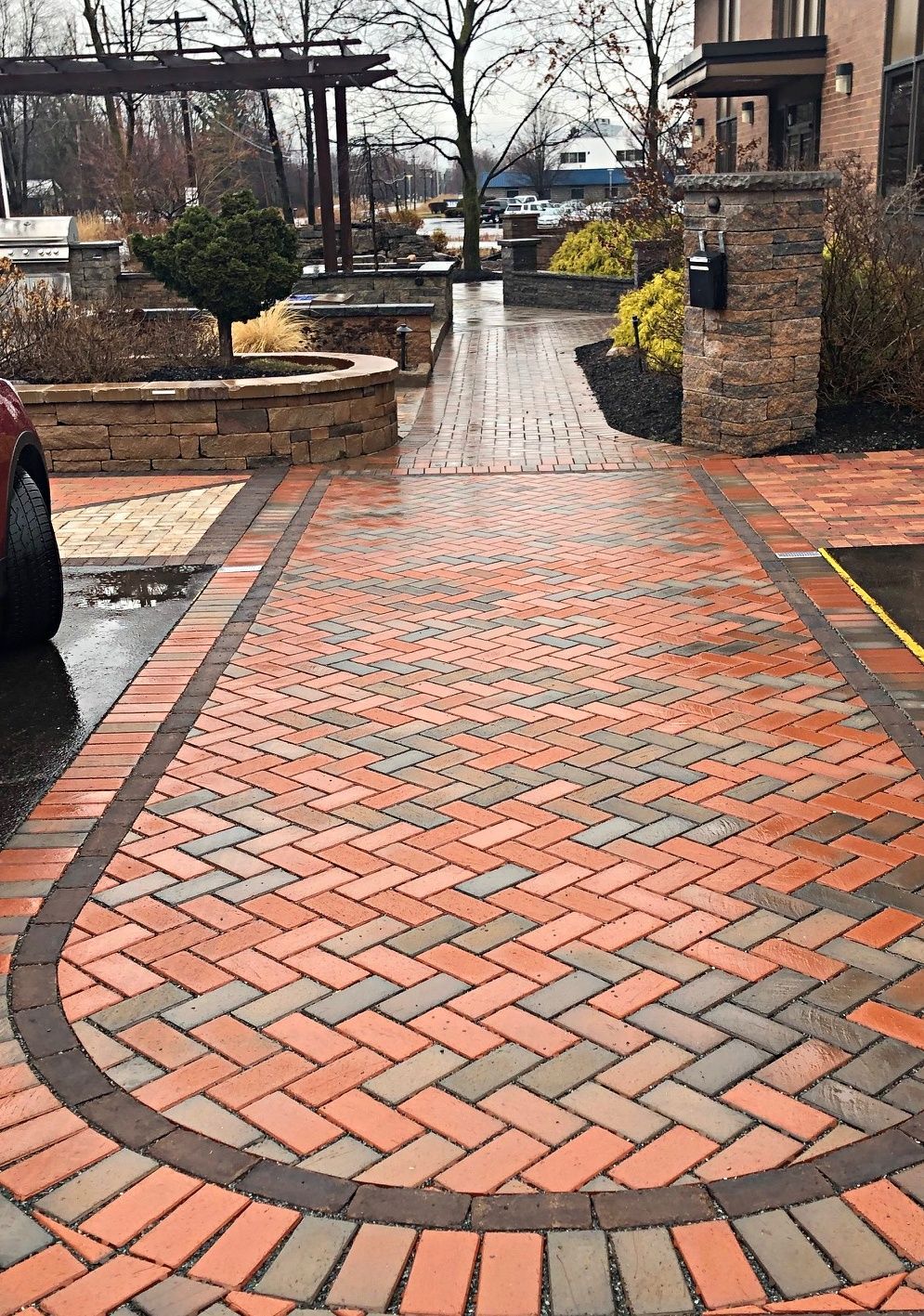 Brick walkway with red and gray patterned pavers, curved edge, and surrounding landscaping.