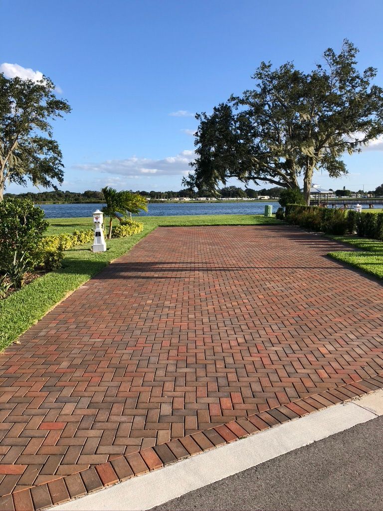 Brick driveway leading to a body of water under a blue sky, with trees and grass bordering.
