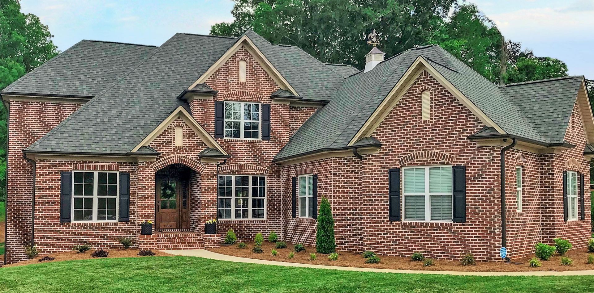 Brick house with dark shutters and a grey roof. Green grass and trees in the background.