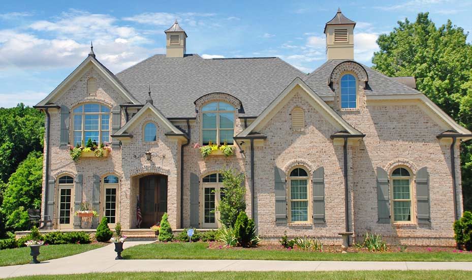 Two-story brick house with gray roof, multiple arched windows, and decorative shutters, on a green lawn.