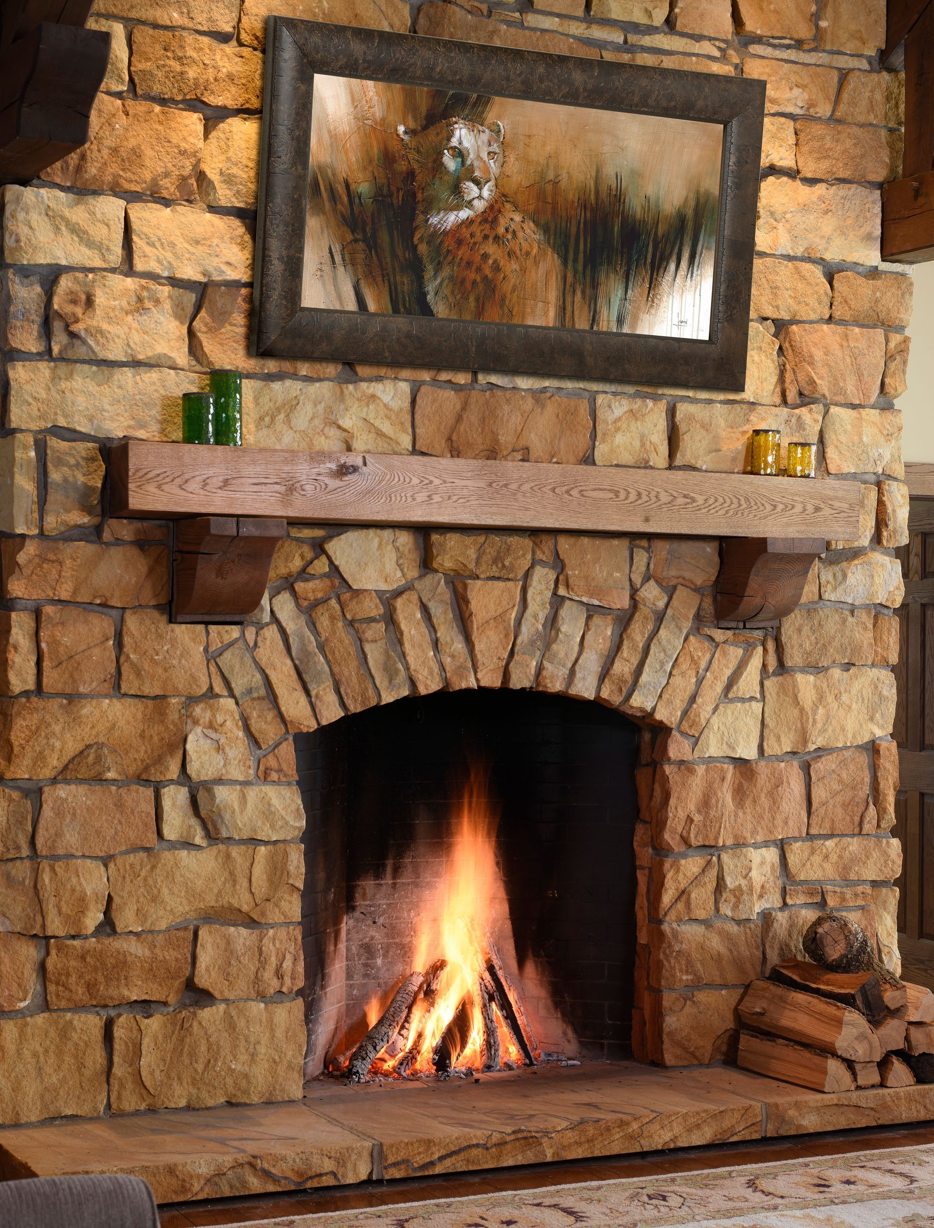 Stone fireplace with arched opening, burning fire, wooden mantel, and framed painting of a wild animal.