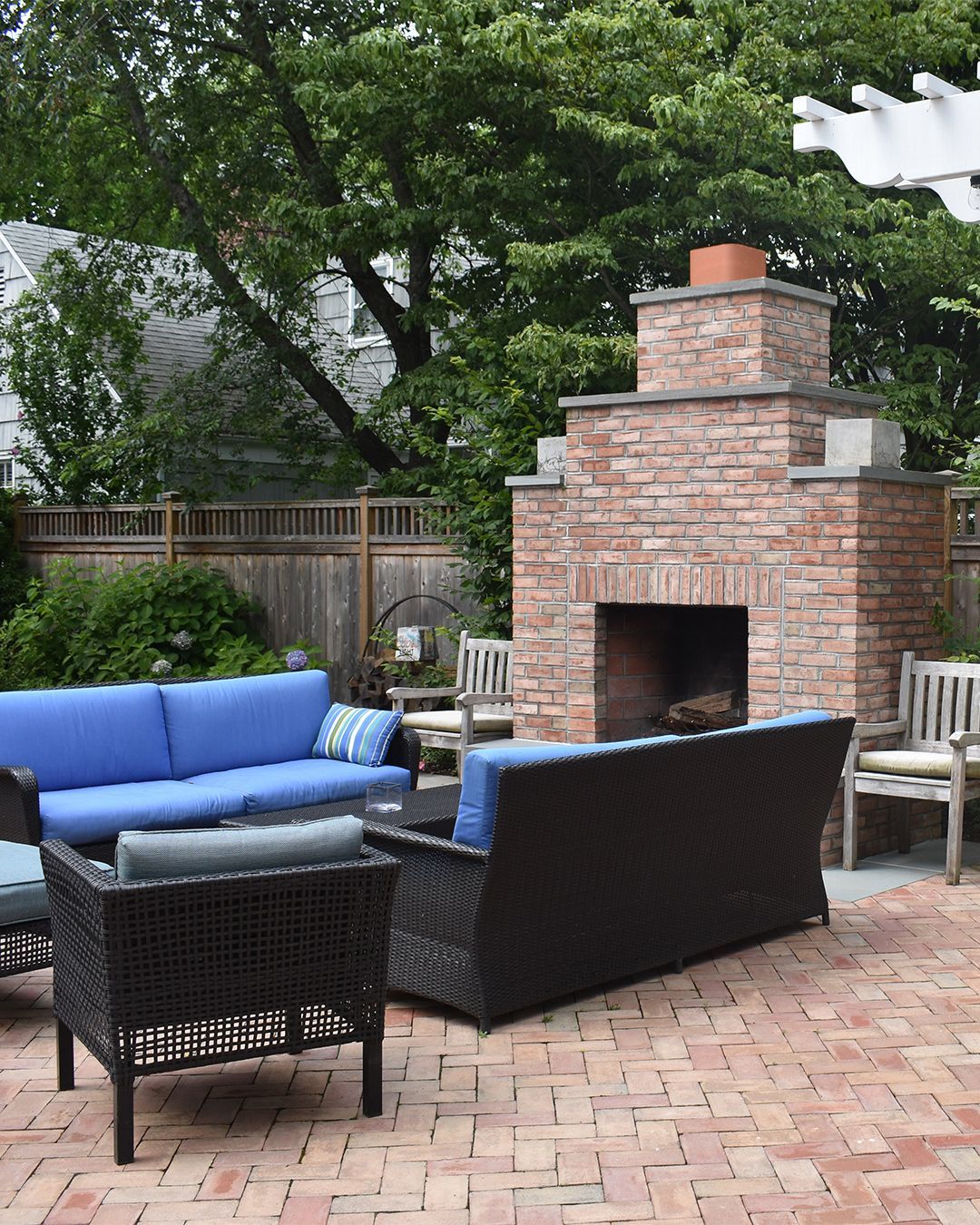 Patio with brick fireplace, blue outdoor furniture on brick pavers, wooden fence, lush greenery.