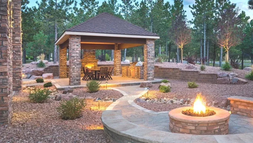 Stone-built gazebo with outdoor kitchen, fire pit, and landscaped yard lit at dusk.