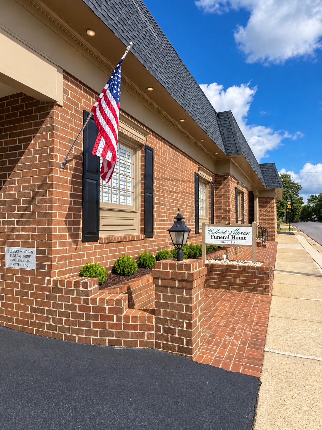 Brick building with dark roof, porch, and a driveway. Trees and blue sky in the background.