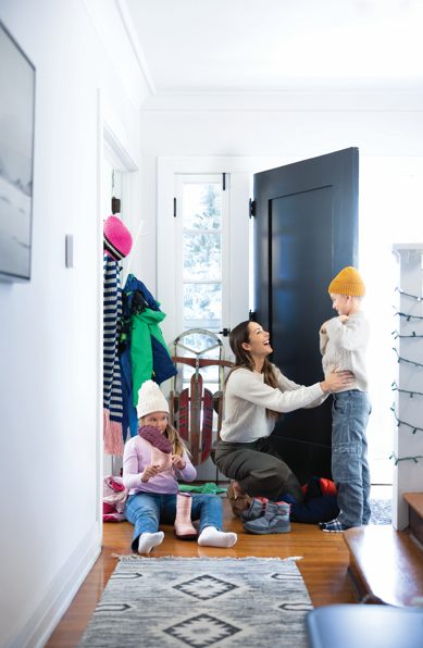 Woman helping a child with a yellow hat in a bright hallway, with another child seated nearby and coats hanging by the door
