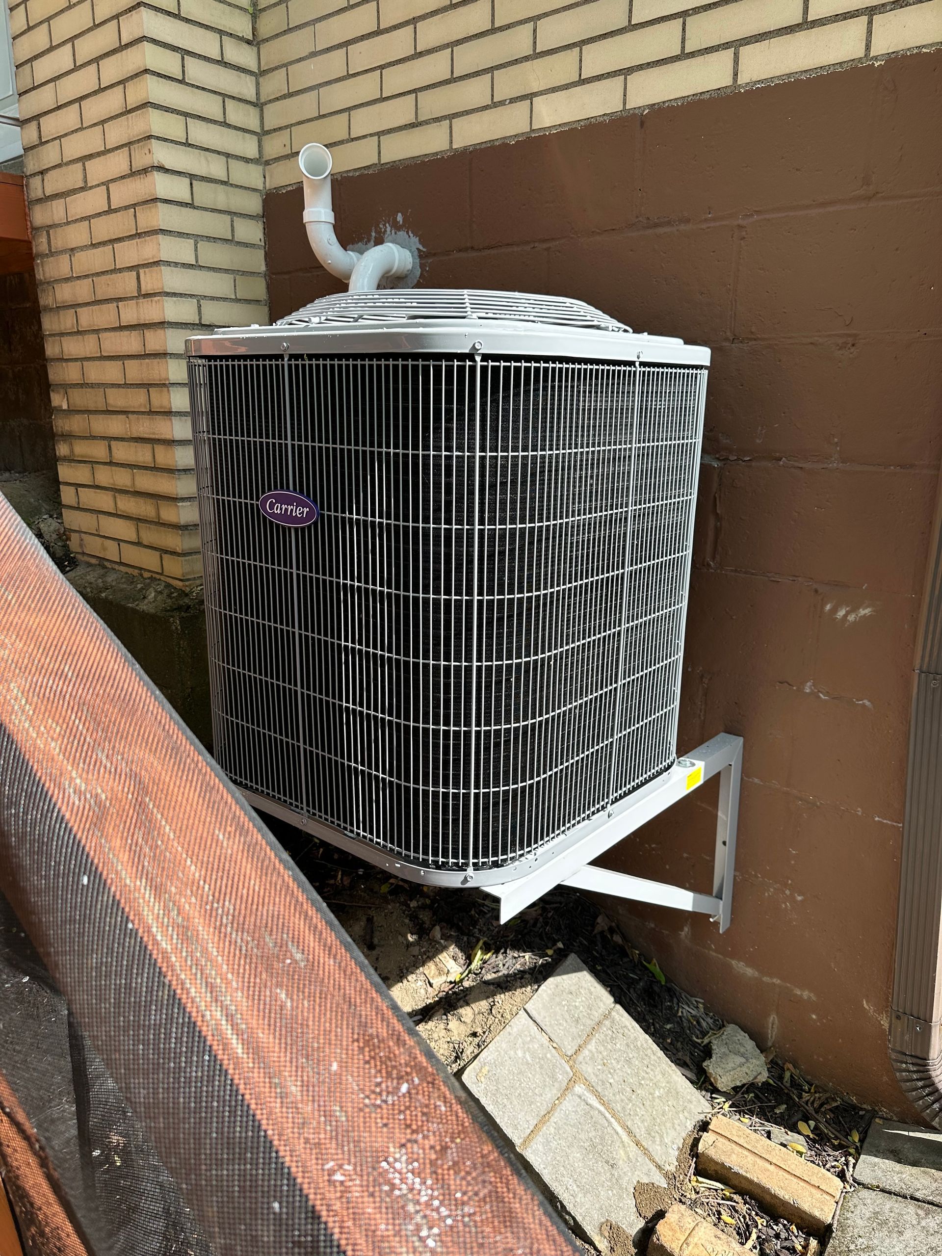 Outdoor air conditioner condenser unit beside a brick wall, with bent metal grilles and debris nearby