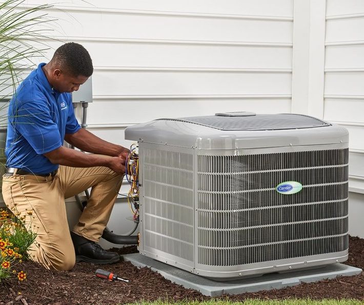 Technician kneeling beside an outdoor air conditioner, servicing the unit near a house wall.
