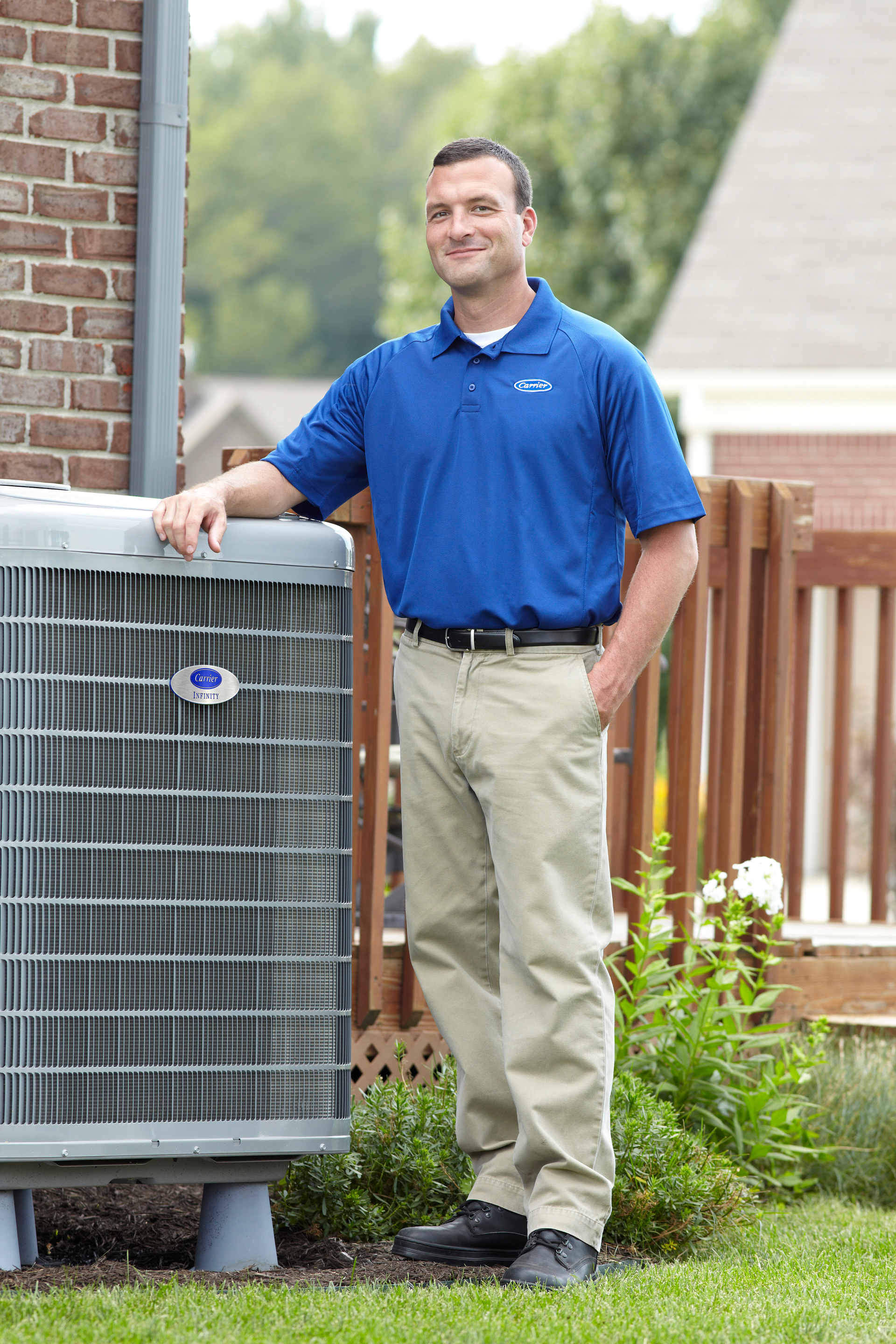 Man standing beside an outdoor HVAC unit, wearing a blue polo and khaki pants, in a yard near a house.