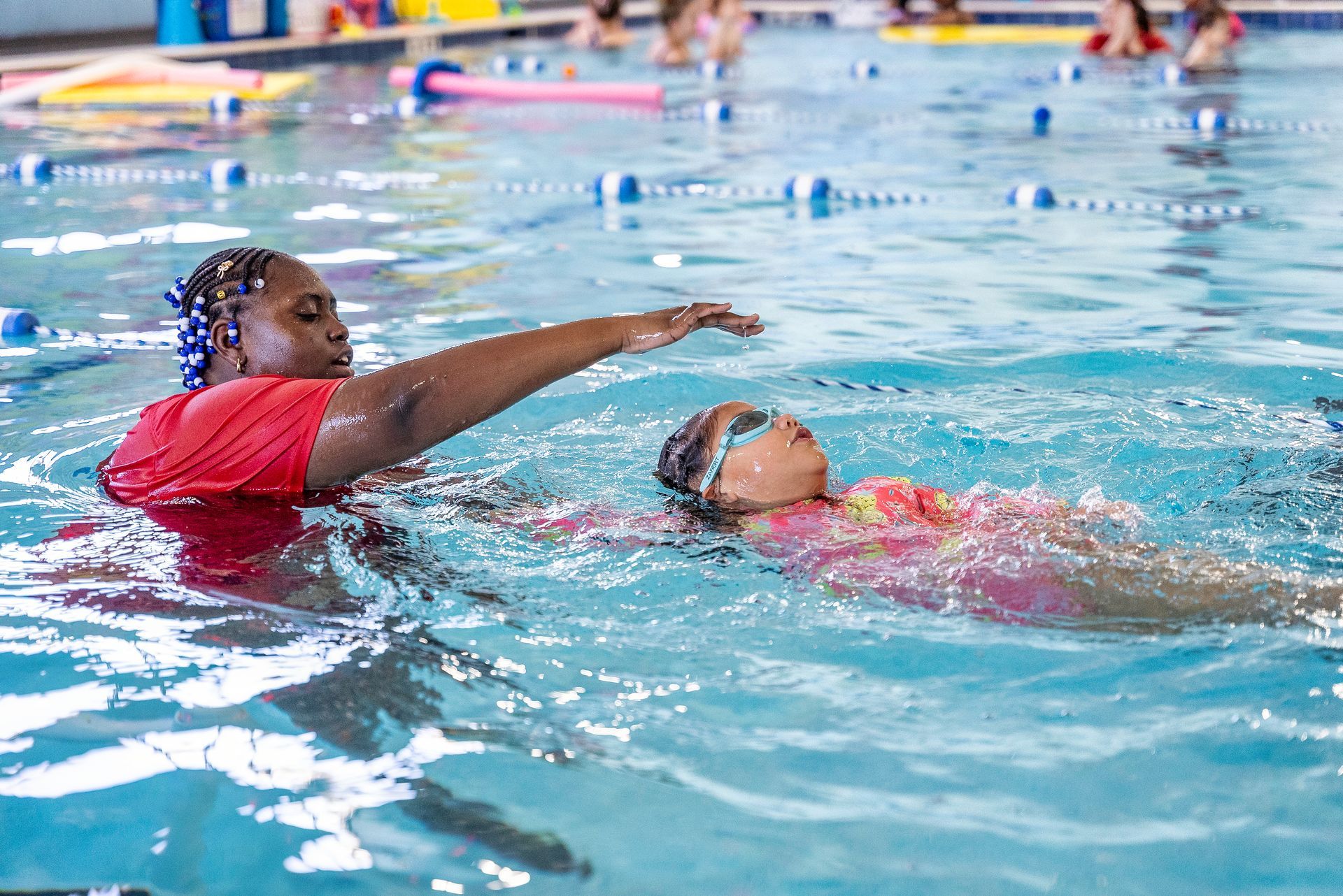Man lifting a child in a pool, water splashing. Child wears swim trunks and goggles, colorful mural background.