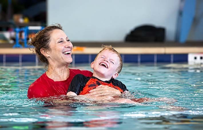 Woman and child laughing in a pool. Woman wears red shirt and holds the child wearing a black and orange rash guard.