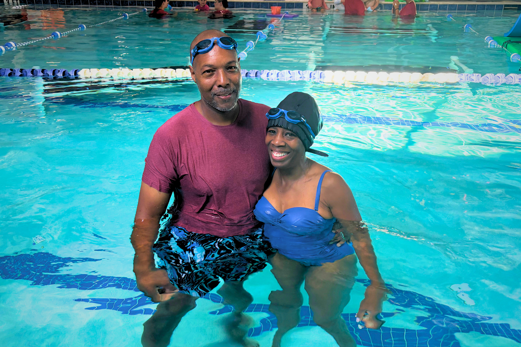 Two children in swimwear at a pool are splashing and laughing. A woman in the water plays with them.
